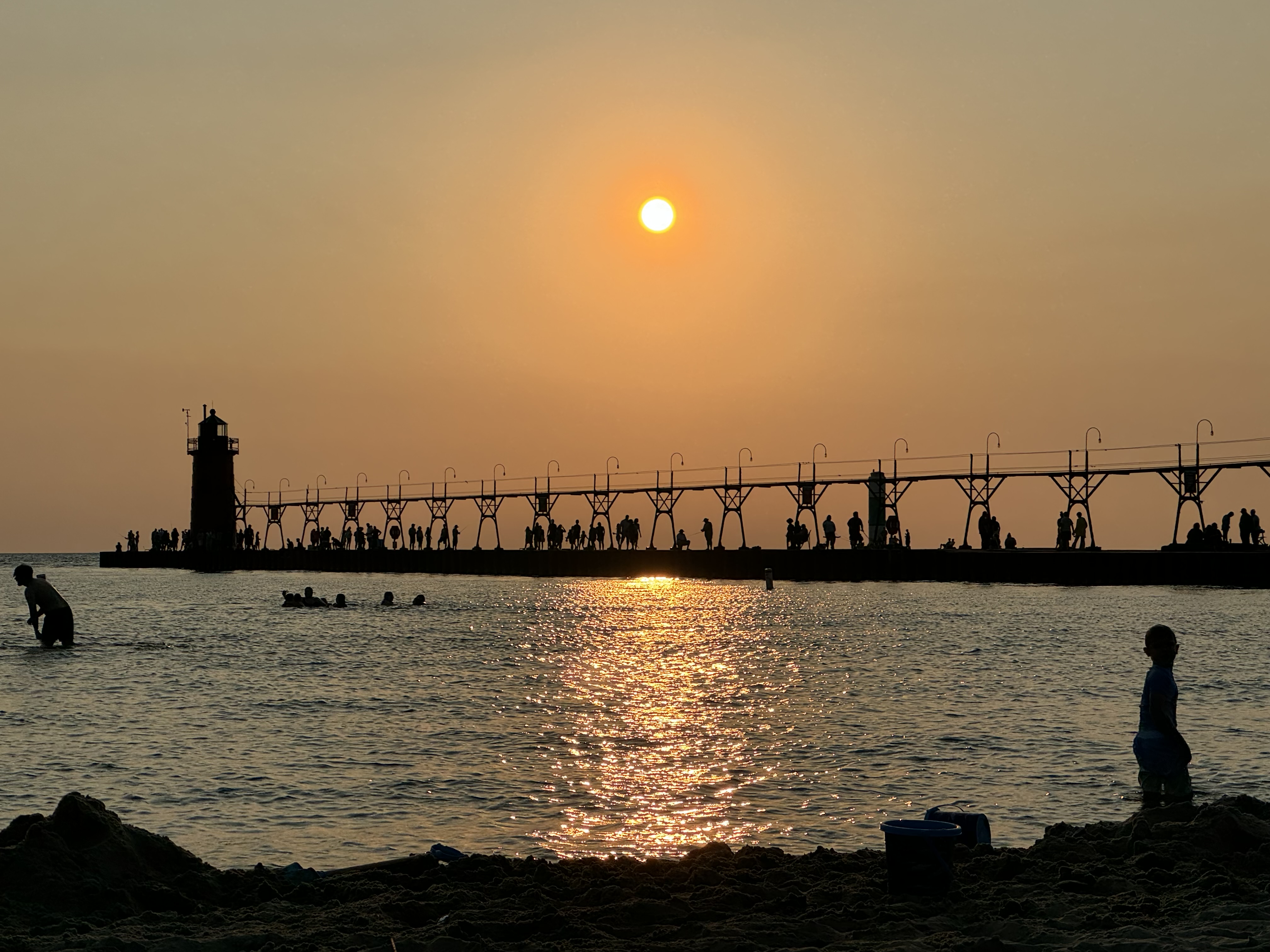 Sunset over the South Haven Lighthouse and pier with silhouettes of people walking along the pier and swimming in Lake Michigan.