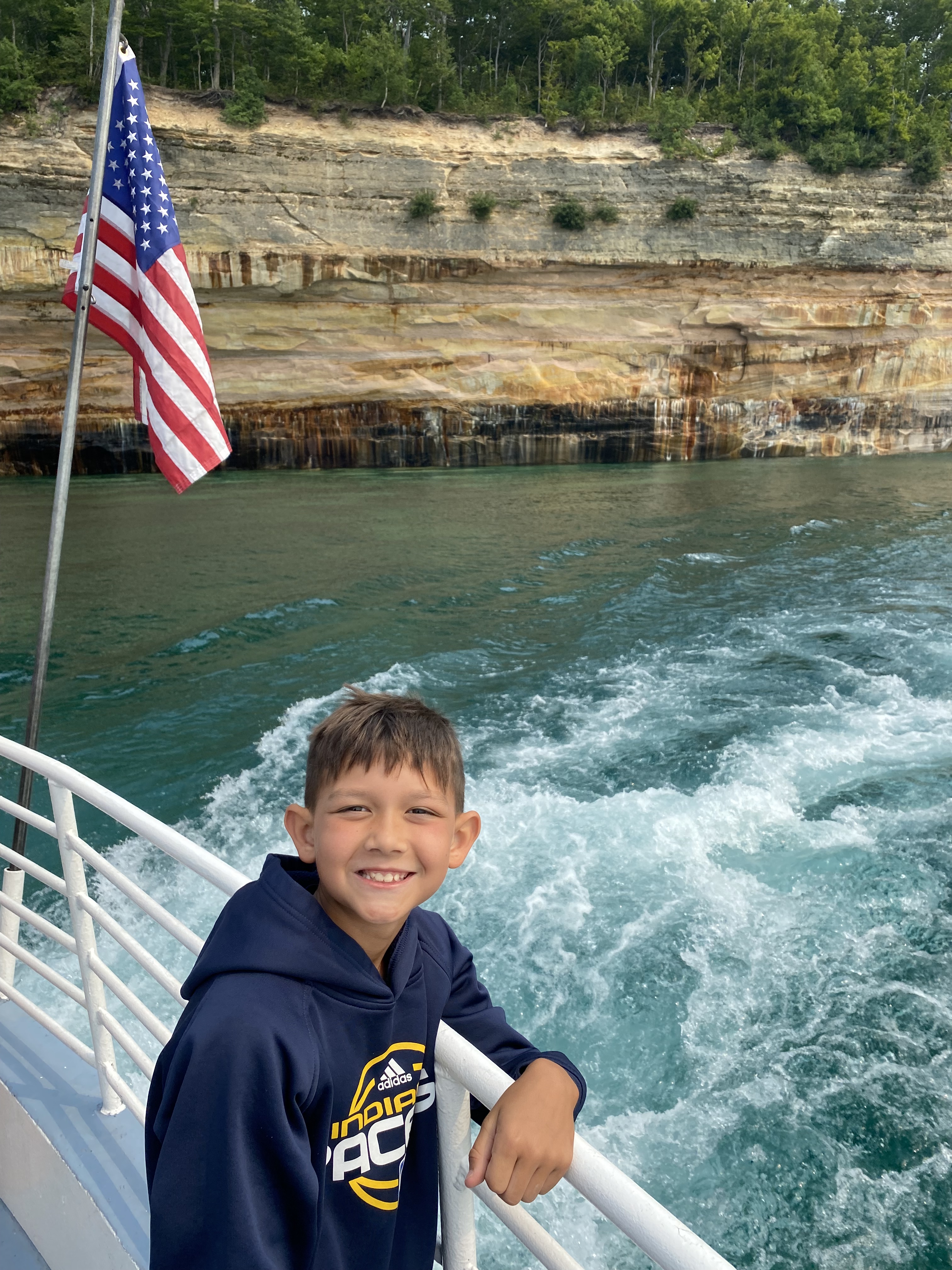 Child smiling on a Pictured Rocks boat tour with an American flag and Lake Superior cliffs behind him