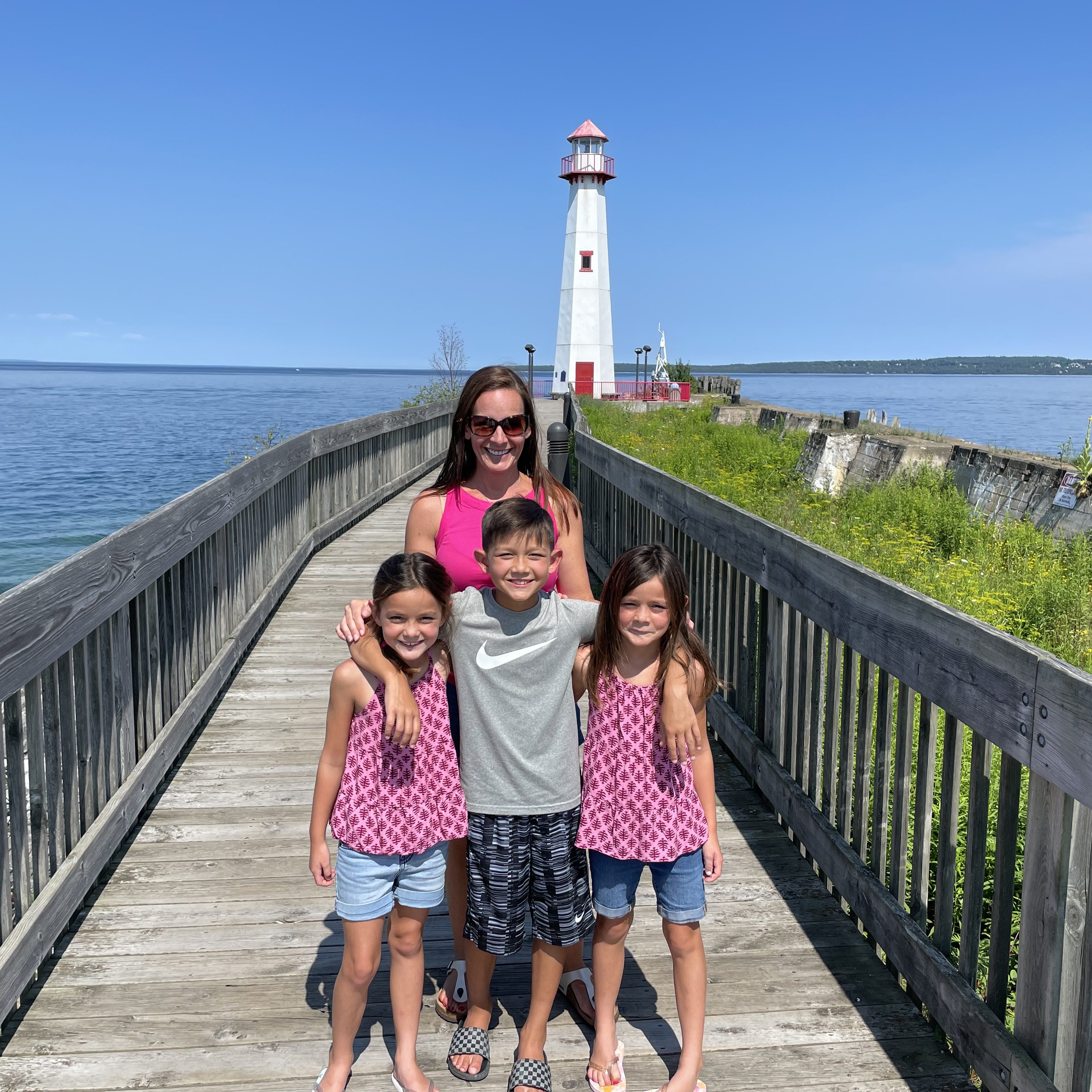 Family of five posing on the boardwalk leading to a white lighthouse at Mackinac Island with blue water and sunny skies in the background.