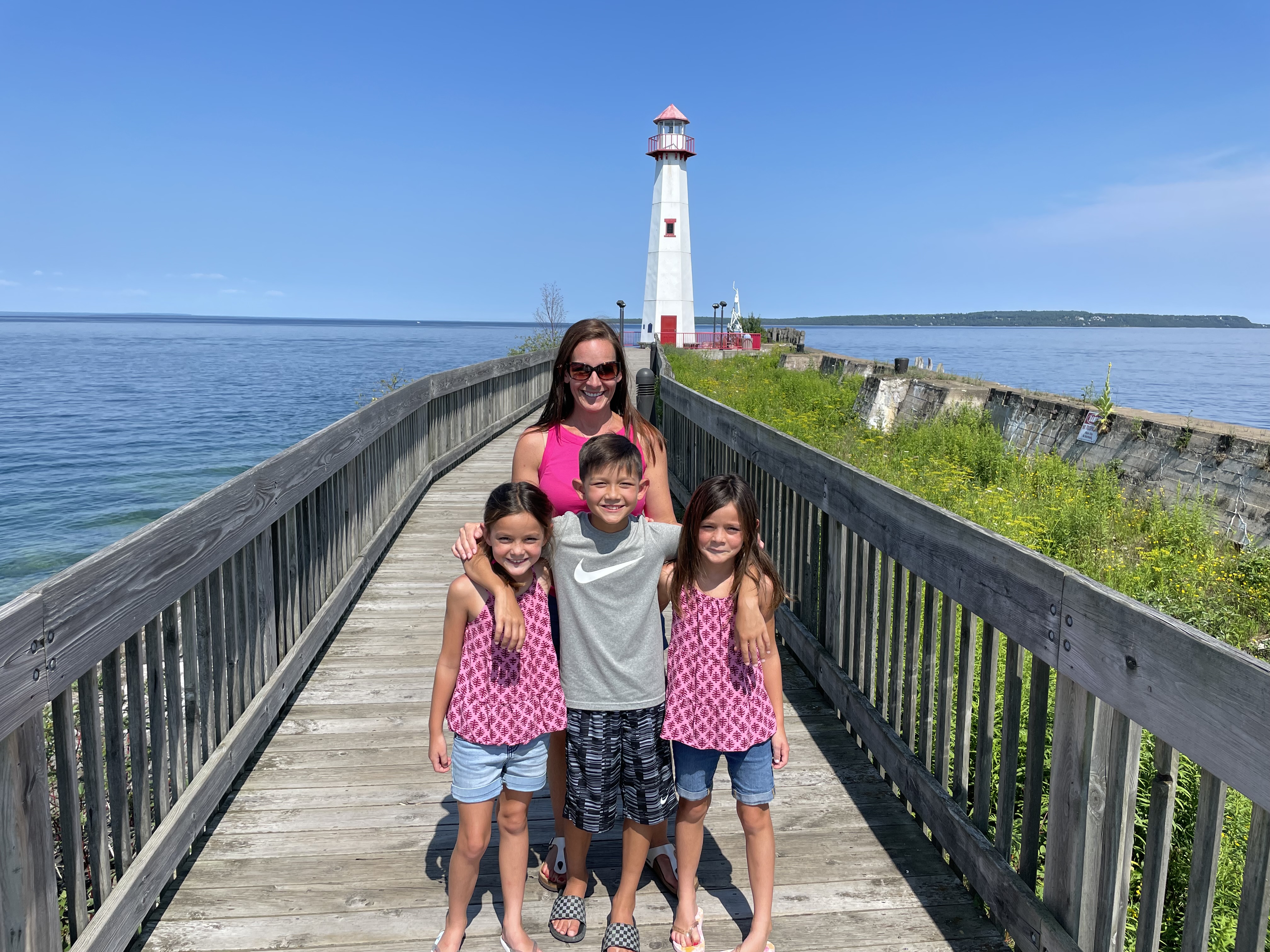 Family of five posing on the boardwalk leading to a white lighthouse at Mackinac Island with blue water and sunny skies in the background.