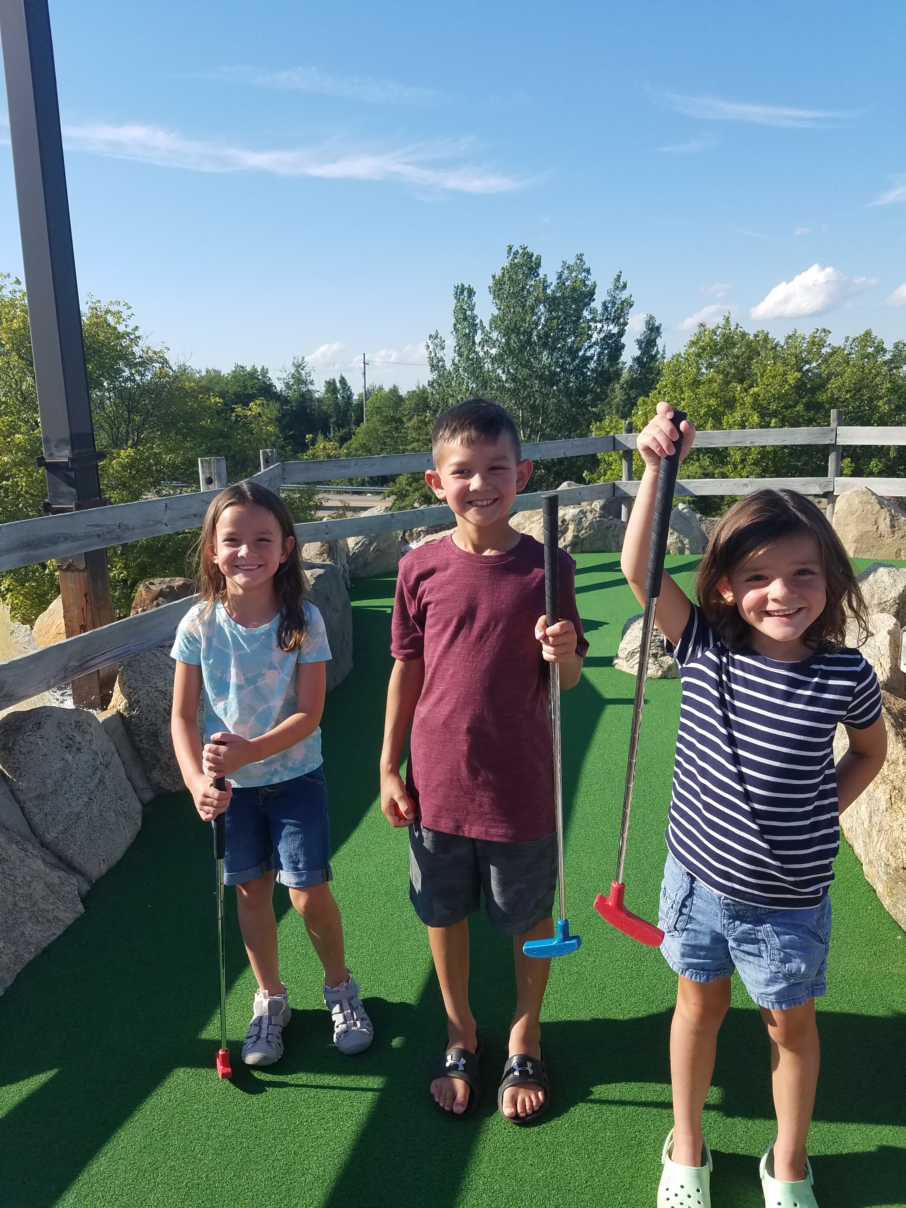 Three kids holding mini golf putters at an outdoor mini golf course in Grand Haven, Michigan.