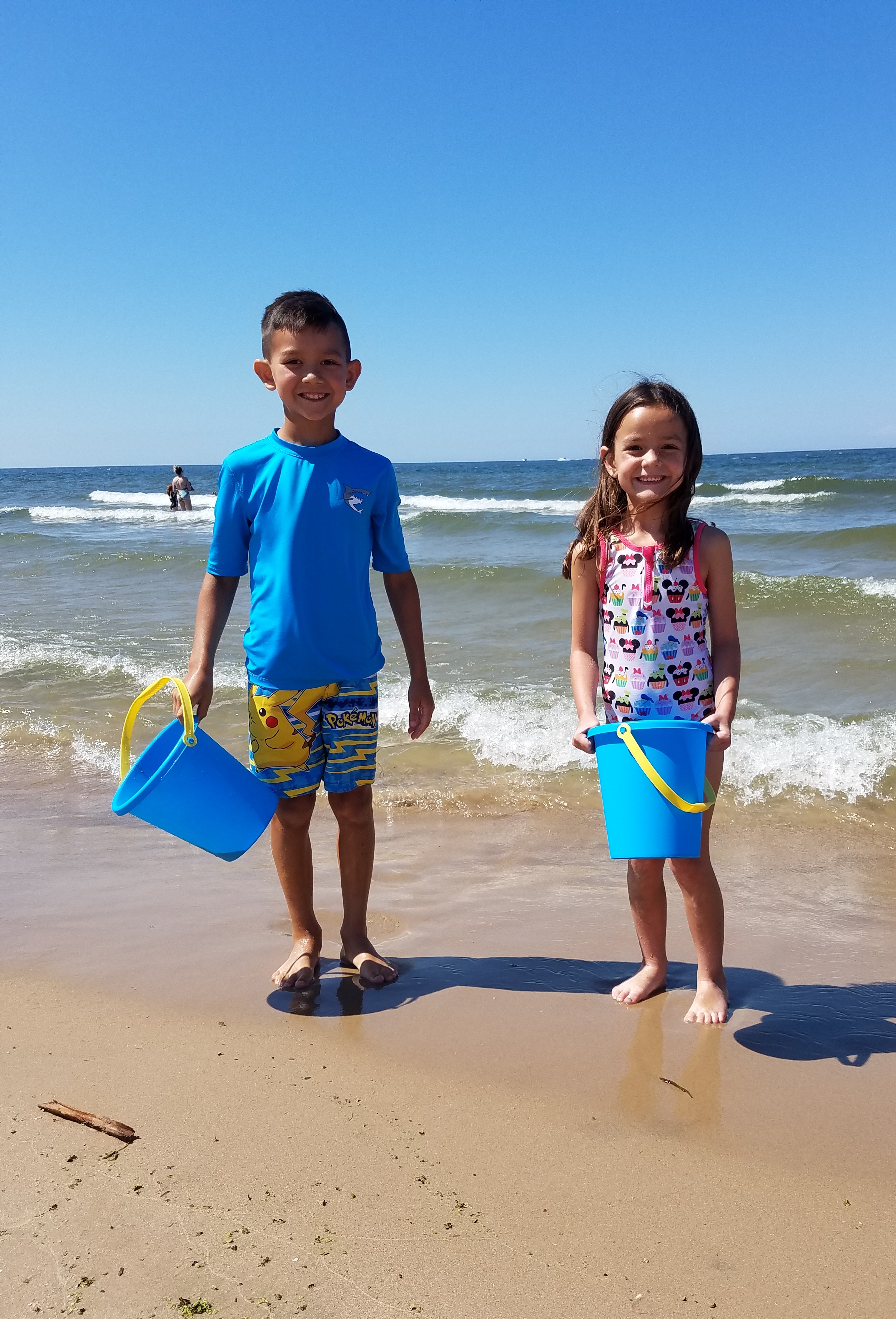 Two children holding blue beach buckets while standing along the shoreline of Lake Michigan in Grand Haven, Michigan.